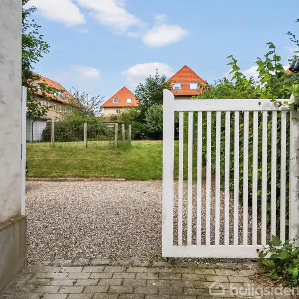 En hvid havelåge står åben, fører ud til en grussti omgivet af grønne buske. I baggrunden ses en græsplæne og rødtagsbygninger under en blå himmel.