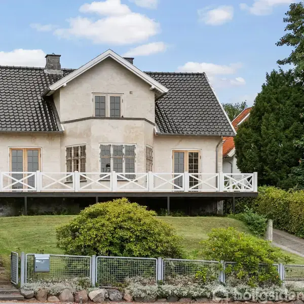 Et hvidt hus med gråt tag står i en velplejet have med græs og buske, omgivet af en hæk og en stenbelagt sti.