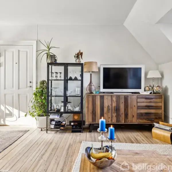 Fladskærms-TV står på en træskænk; lampe og planter omkranser det. Stue med trægulv, blå sofa og glasvitrine. Dekorative genstande på skænk; lys og skål på bord.