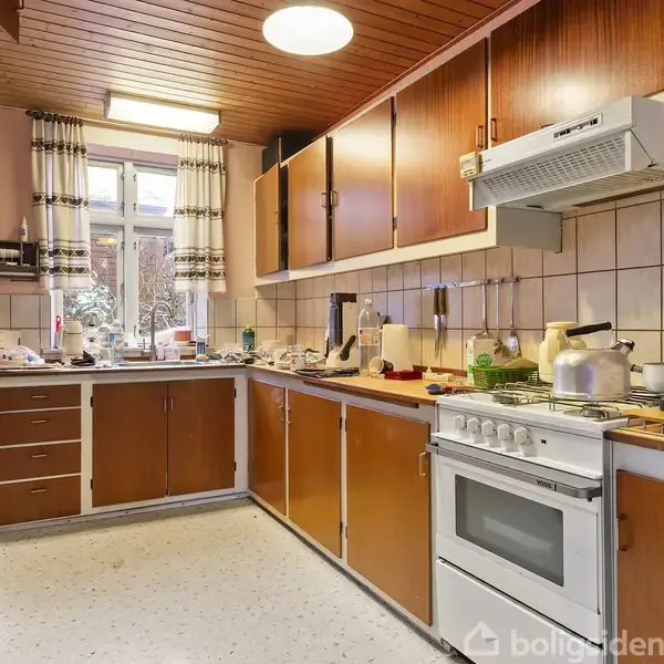 Køkken med brune skabe fyldt med køkkenudstyr og porcelæn, hvor en gryde simrer på komfuret; træloft og vindue med gardiner skaber en hyggelig stemning. Ingen tekst til stede.