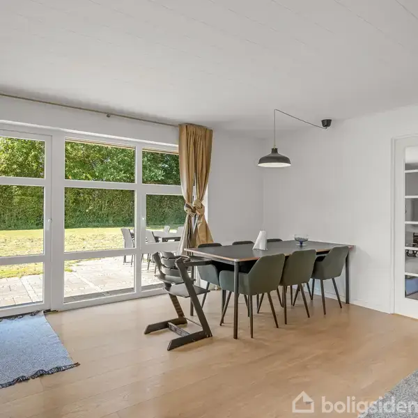 Spisestue med stort spisebord og seks stole, placeret ved glasdøre, der åbner ud til en terrasse med udsigt til have. Lampen hænger over bordet, skaber hyggelig atmosfære.