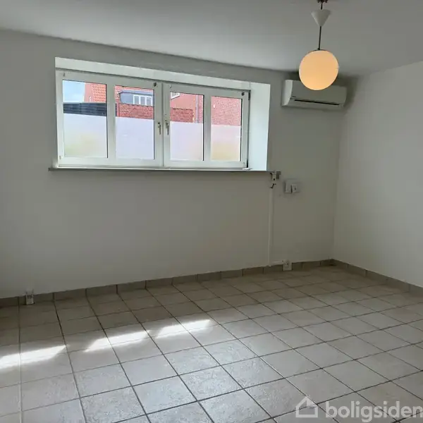 Et tomt værelse med flisegulv, en rund loftlampe, et lille vindue med udsigt til en murstensbygning og en vægmonteret airconditionenhed, der skaber en lys og ren atmosfære.