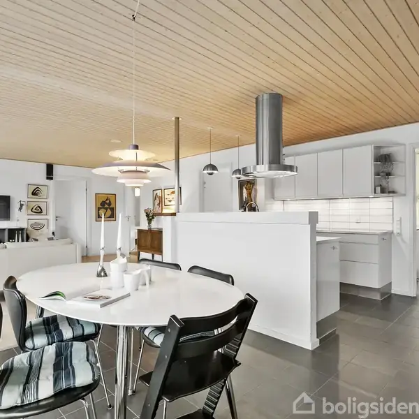 Et moderne, åbent køkken-alrum med spisebord og stole foran, køkken med hvide skabe og stålventilation til højre, stue med tv og sofa i baggrunden, træloft og flisegulve.