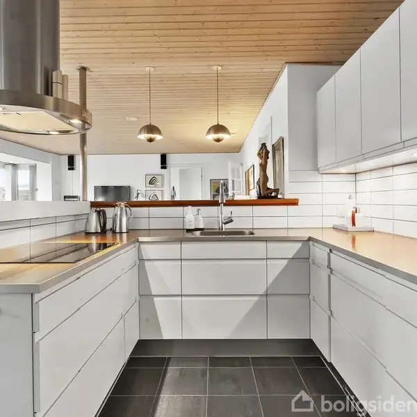 Et moderne køkken med hvide skabsfronter udgør hovedfokus. En marmorplade ligger ovenpå, og der er installeret to hængende lamper. Rummet har flisegulv og træloft i et lyst miljø.