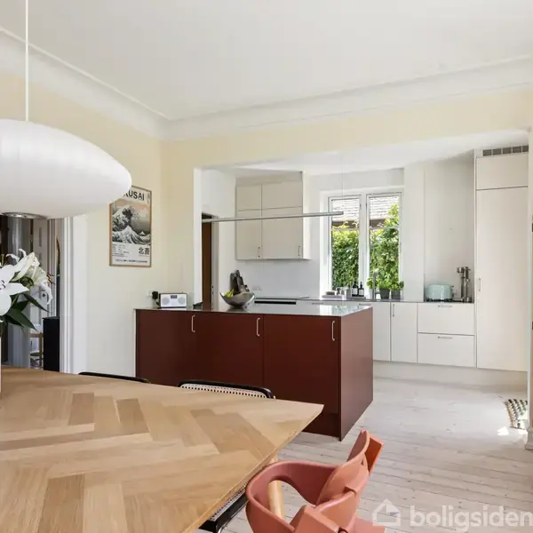 Et moderne køkken-alrum dominerer billedet med et træbord, en hvid pendellampe og blomster. I baggrunden ses et åbent køkken med hvide skabe og en stor ø.