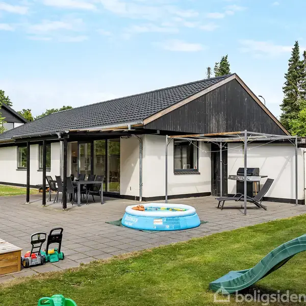 En hvid bungalow med sort tag står på en flisebelagt terrasse. Omkring huset er der græsplæne, en oppustelig pool, et legetårn med rutsjebane og havemøbler.