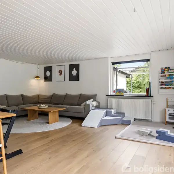 En stue domineret af en stor grå sofa placeret på et rundt tæppe. Den har træbord foran, legetøjstrappe ved vinduet, og billeder på væggen. Trægulve skaber en hyggelig atmosfære.