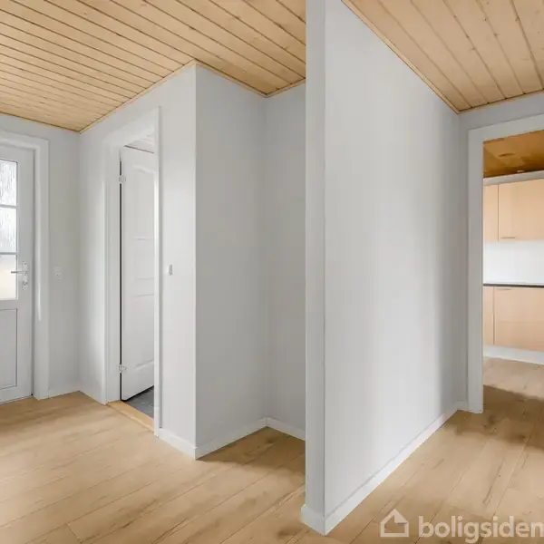 En lys gang med trægulv og træloft fører ud til en dør med glasruder. Til højre ses en åbning ind til køkkenet med lyse skabe.