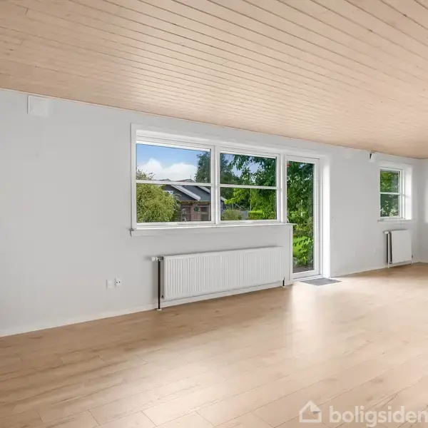 Et lyst, tomt rum med trægulv og træloft indeholder en rød skammel. Store vinduer giver udsigt til træer og et hus. En radiator er monteret under vinduerne.
