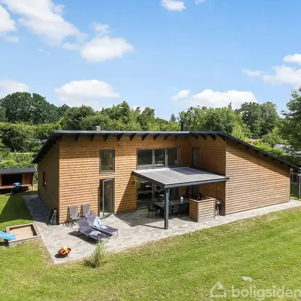 Træhus med fladt tag står på en græsplæne, omgivet af træer. En solterrasse med liggestole ses foran huset. En rutsjebane er placeret i venstre side.