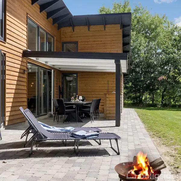 Liggestol står på terrasse, hvor en bålgryde brænder. Rundt om et bord står stole. Huset er træbeklædt med store vinduer. Græsplæne og træer i baggrunden under en blå himmel.