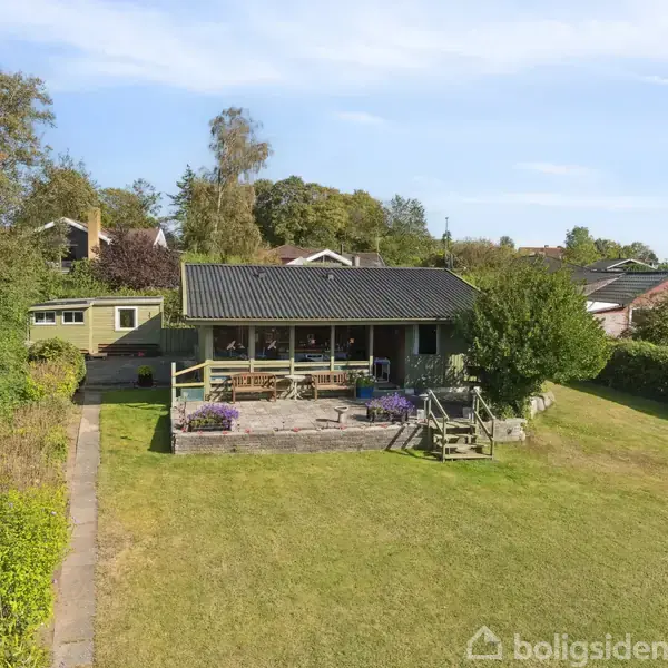 Hus med sort tag står på stor grøn græsplæne. Der er en veranda foran huset med blomster rundt. Omgivet af træer og buske, beliggende i et boligområde.