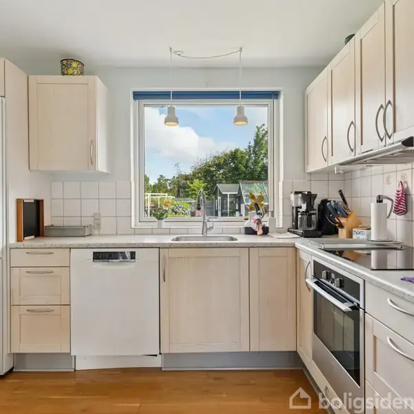 Køkken med lyse træskabslåger og hvidevarer, herunder køleskab med magneter, opvaskemaskine, og ovn. Et vindue med udsigt til en have. Bordpladerne har diverse køkkenredskaber.