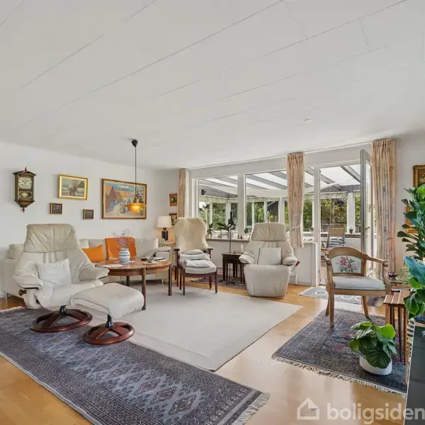 Stue med lyse lænestole arrangeret omkring et sofabord. Store vinduer giver udsigt til haven. Rummelige tæpper dækker trægulvet; planter og vægkunst skaber en hyggelig atmosfære.