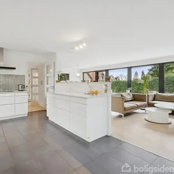 Køkken-alrum har hvide skabe, stående rustik emhætte, og stor sofa. Glasdøre og store vinduer åbner mod en frodig have. Skakternet gulv i køkkenet og lyst trægulv i stuen.