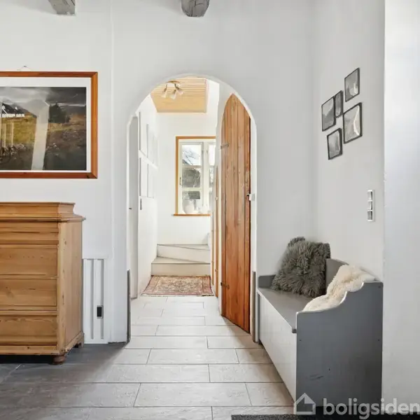 Et lyst rum med en trædør i forgrunden, en buegang i midten, og en kommode til venstre med en lysestage ovenpå. Flere indrammede billeder hænger på væggen.