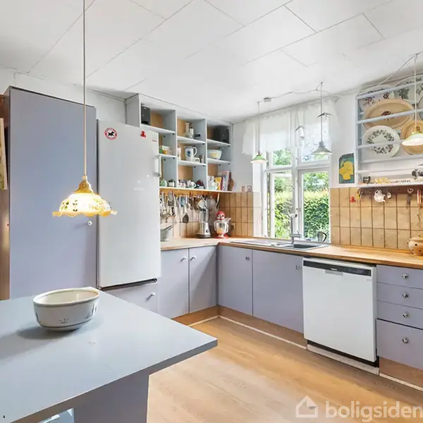 Køkken med lyseblå skabe, hvid opvaskemaskine og køleskab. En skål står på en lille bordplade, og over den hænger en lampe. Vinduet har udsigt til grønne buske.