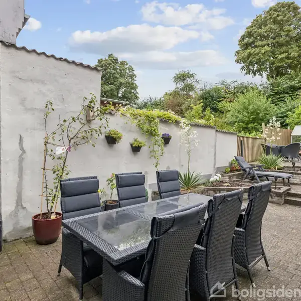 Udendørs spisebord med otte sorte kurvestole står på en terrasse af brosten. En hvidmalet mur med blomster hænger ved siden af. Træer og buske omkranser området i baggrunden.