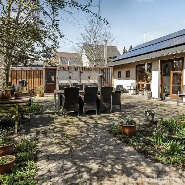 Terrasse med bord og stole i centrum omgivet af blomsterkrukker. En pergola og et hus med solpaneler i baggrunden skaber en hyggelig haveatmosfære.