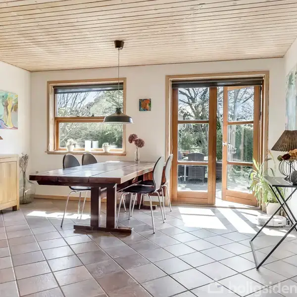 Spisebord med stole står i lyst rum med flisegulv. Store vinduer og glasdøre giver udsigt til terrasse og have. Træloft og dekorationer skaber en hyggelig atmosfære.