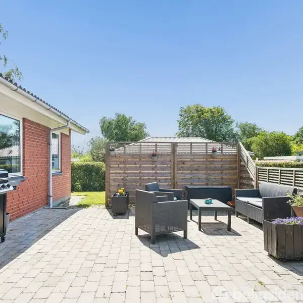 Terrasse med flisebelægning, møbleret med sort sofa, stole og bord. En grill står ved huset med rød murstensvæg. Træplantekasser og træhegn omgiver området. Klar blå himmel.