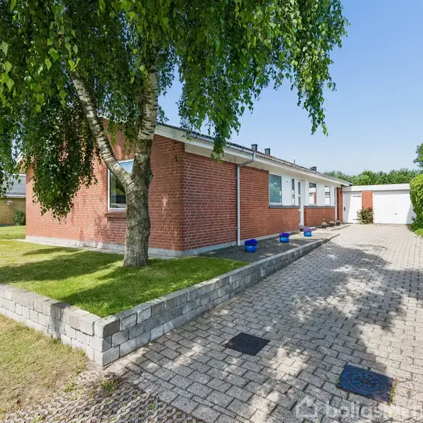 En rød murstenshus med fladt tag står ved siden af en brolagt indkørsel. En stor træ i forhaven giver skygge. Området er grønt og solrigt.