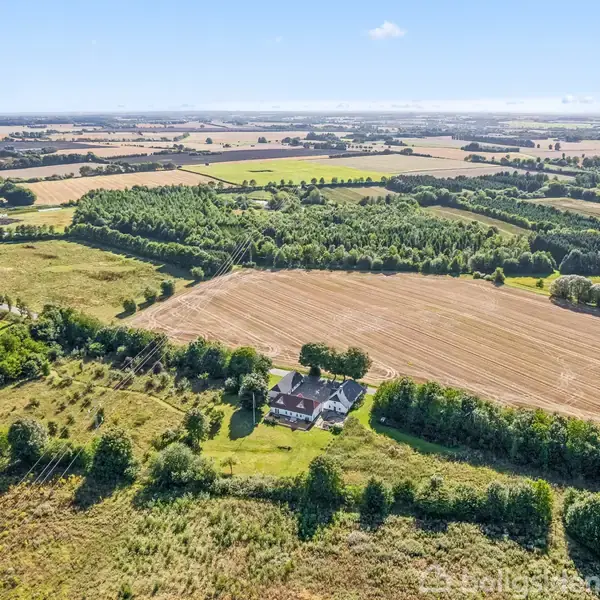 Et hus ligger midt i et åbent landskab omgivet af marker og træer. Landskabet strækker sig til horisonten under en blå himmel.