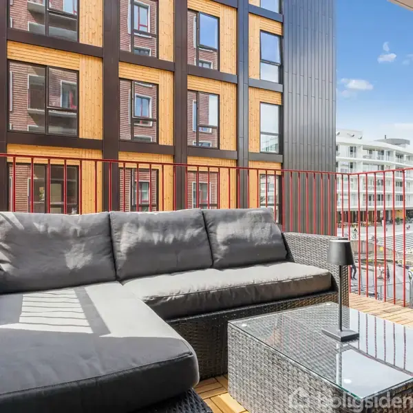 En grå hjørnesofa står på en altan med glasbord foran. Bordet har en lille lampe. I baggrunden ses et moderne lejlighedskompleks med farverige detaljer og en klar blå himmel.