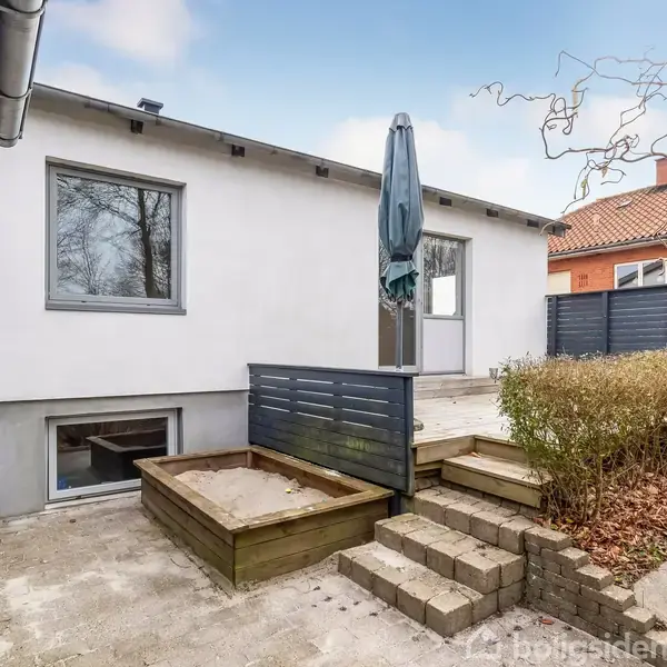 Husets terrasse indeholder en sandkasse og en parasol. Trapper fører op til en hvidmalet murstensbygning. Omgivet af beplantning og et hegn i et boligområde.