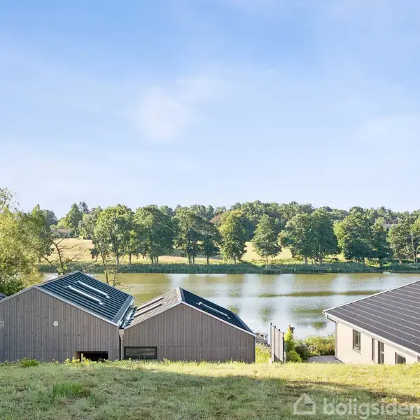 Huse med moderne tagkonstruktion står på en grøn bakkeskråning. Baggrund viser en sø omgivet af tæt skov. Himlen er klar og blå.