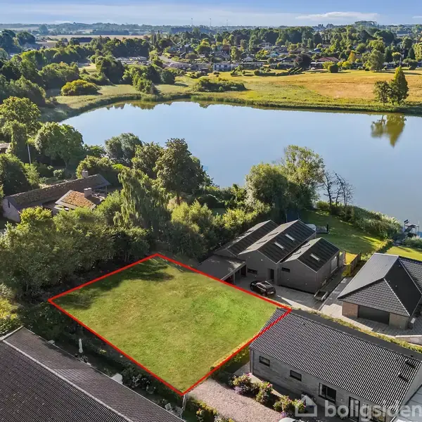 En tom grund omkranset af et rødt omrids ligger mellem huse ved en sø. Omkring er grøn vegetation og flere huse.