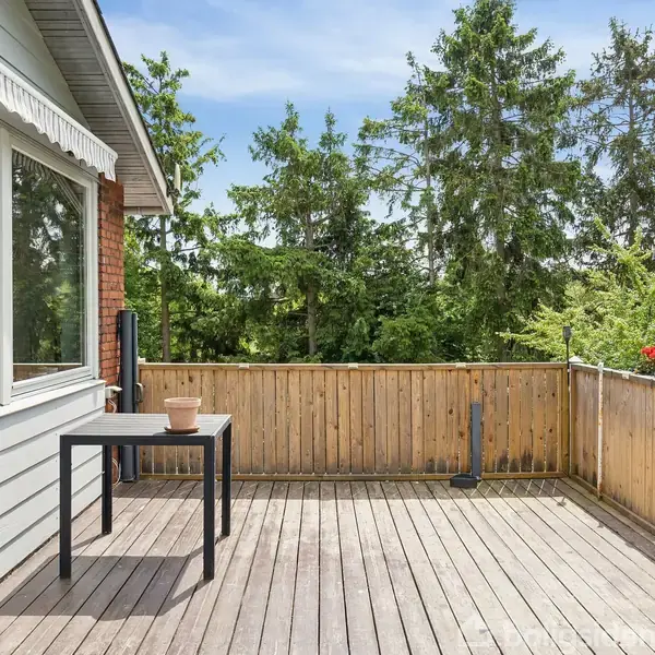 Et træbord med en terracotta potte står på en terrasse omkranset af et hegn. Grill står til højre, omgivet af frodig skov.