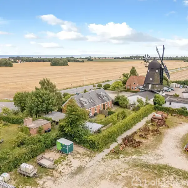 Vindmølle står stille ved siden af en gård med murstensbygninger. Omkringliggende marker strækker sig mod horisonten. Landbrugsmaskiner og containere er spredt i forgrunden.