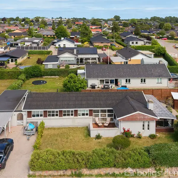 En række enfamiliehuse med velholdte haver ligger i et boligområde. Biler er parkeret i indkørsler. Græsplæner og grønne områder omgiver husene. Himlen er blå med få skyer.
