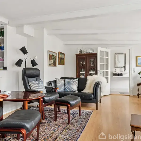 En stue med sorte lædermøbler og et sofabord står på et orientalsk tæppe. En skænk med planter og et vitrineskab ses i baggrunden ved siden af et vitrineskab.