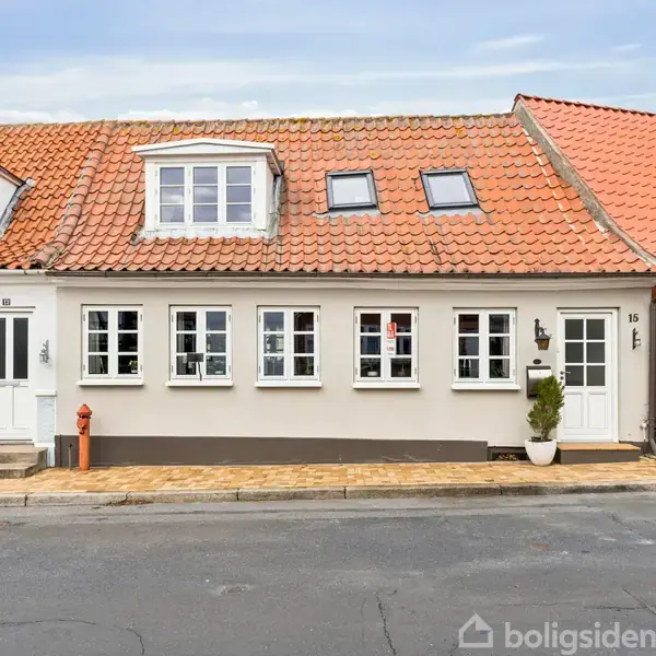 Rækkehus med orange tegltag og hvide vinduer står stille i en byvej. Huset har skodder, og døren er placeret ved siden af en potteplante. Husnummeret "13" er synligt.