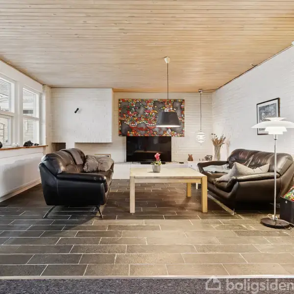 Stue med sorte lædersofaer omkring et lyst sofabord, farverig kunst på en hvid væg, glasskab med dekorationer til højre og store vinduer til venstre.