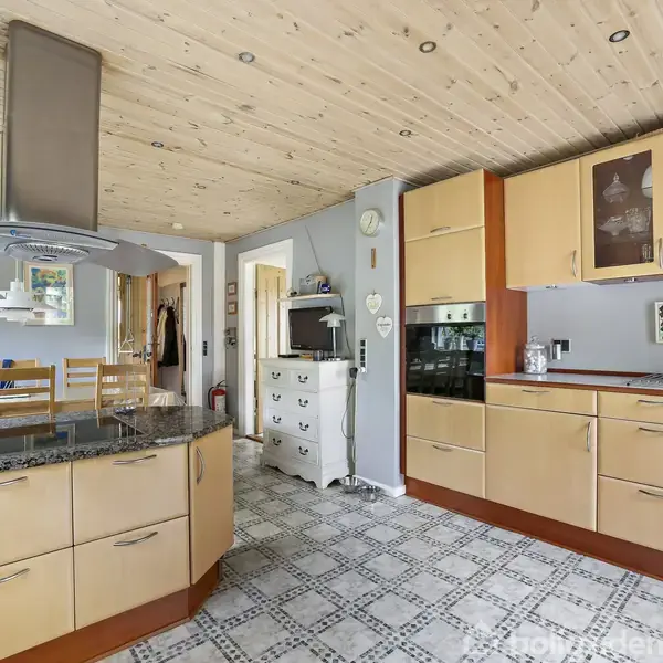 Et lyst køkken med træloft har et centralt køkkenø med granitbordplade, stolerække og emhætte. På højre væg er der lyse træskabe, glasvitrineskabe, indbygget ovn og en væghylde.