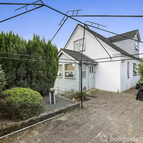 Et hus med hvid facade og sort tag står omkranset af træer og buske på en flisebelagt terrasse. En tørrestativstruktur strækker sig over terrassen, hvor der også er et bord med stole og en grill.
