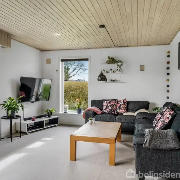 En stue med grå sofa, dekoreret med blomstrede puder, står ved et stort vindue. Et træbord foran sofaen og en vægmonteret TV-skærm på venstre side supplerer indretningen.