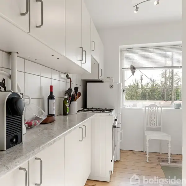 Køkkenbord med kaffemaskine og flasker står langs en væg med hvide skabe. I baggrunden er der et vindue og en stol ved et lille spiseområde. Ingen tekst.
