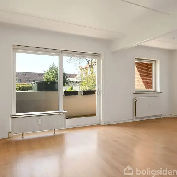 Rummet har trægulv, store vinduer og en dør, der åbner ud til en balkon. I baggrunden ses en have med træer og buske samt en murstensmur.