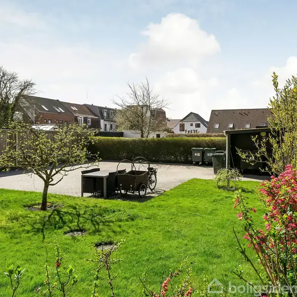 Grønt græsareal med et lille træ i midten, omgivet af buske og blomster. Til venstre står havestole og borde. I baggrunden ses huse og en carport.
