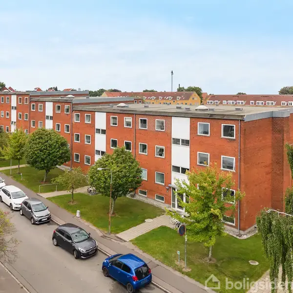 Røde murstensbygninger med flade tage står langs en vej med parkerede biler. Træer og græsplæner omkranser bygningerne i et boligområde.