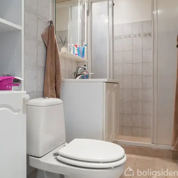Toilet med hvidt skab og hylder til venstre indeholder toiletartikler; brusekabine med glasdør til højre; vask med spejl over. Badeværelset er lyst og funktionelt.