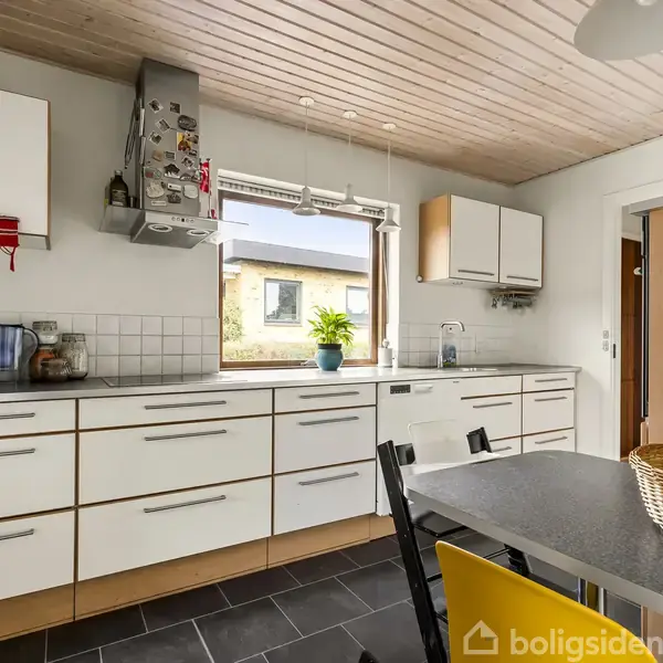 Et moderne køkken har hvide skabe og en sort bordplade. En frugtkurv står på bordet, og et vindue med planter giver udsigt til en gul bygning.