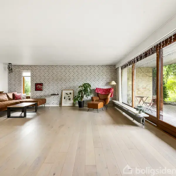 Stue med lyse trægulve og hvide murstensvægge, indrettet med brun lædersofa, stol, kunstværker, planter og stort vinduesparti, der åbner op til en terrasse med grønt landskab udenfor.