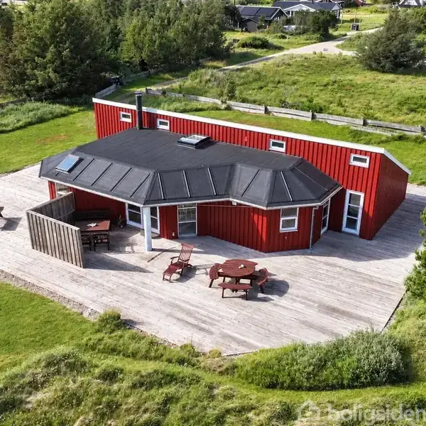 Græsmark med sti fører til hus med terrasse. Solen skinner gennem skyer i en stille, landlig atmosfære. Terrassebord og stole står klar til brug.