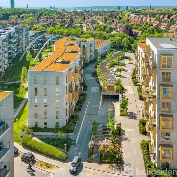 Moderne lejlighedsbygninger står omkring en central, smal gade. Grønne tage præger bygningerne, mens træer og grøn beplantning ses i området. Cyklister og fodgængere færdes i et boligområde med udsigt til natur.