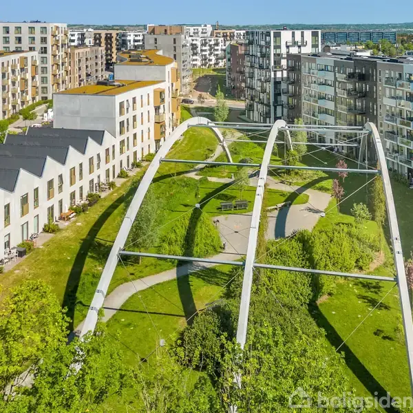 Et boligområde med moderne bygninger omgiver en grøn park. Buen strukturer strækker sig over parken, som indeholder stier og buske. Omkringliggende bebyggelse indikerer en bymæssig kontekst.